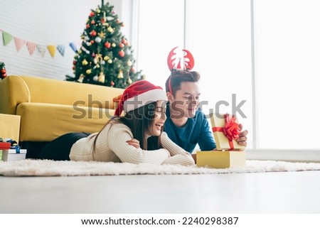 Young happy Asian woman wearing a Santa Claus hat with her boyfriend with a Christmas gift while lying down on the carpet with a Christmas tree in the background. Photo with copy space.
