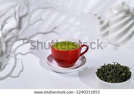 A red cup containing green tea, on a white background with white teapot and a bowl full of dried green tea leaves. 