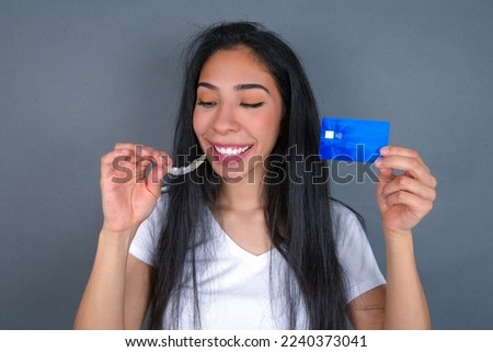 Young beautiful hispanic woman wearing white t-shirt over gray studio background, holding an invisible aligner on one hand and a credit card on the other hand. 