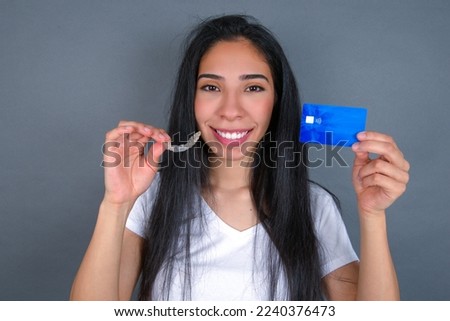 Young beautiful hispanic woman wearing white t-shirt over gray studio background, holding an invisible aligner on one hand and a credit card on the other hand. 