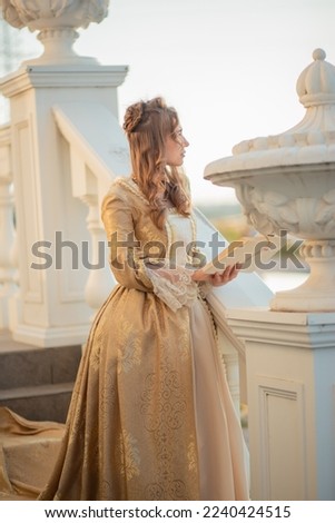 A beautiful young woman in a historic eighteenth century gold dress stands on the stairs of the mansion. Princess in the palace. fabulous image Royalty-Free Stock Photo #2240424515