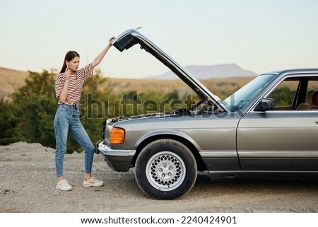 A woman stands outside a broken-down, dangerous old car with the hood open with a wrench on a road trip alone Royalty-Free Stock Photo #2240424901