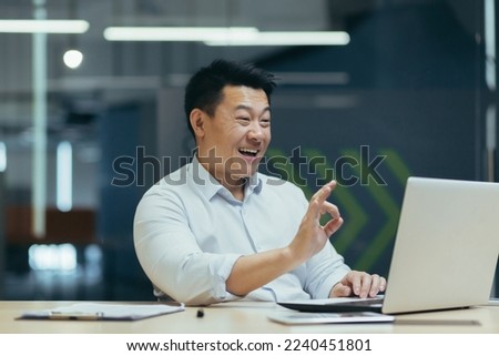 A young Asian businessman conducts an online interview for a job in company. He sits in the office at a table with a laptop, talks through a video call, points with his fingers ok, smiles. Royalty-Free Stock Photo #2240451801