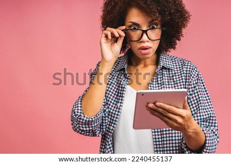 Amazed shocked american woman with curly african hair holding digital tablet and smiling standing over isolated pink background with copy space for text, logo or advertising.