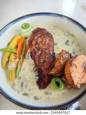 Roasted chicken, potato wedges, carrots, green beans, baby corn, cucumber garnish, and mushroom sauce are arranged beautifully and served on a white plate.  Flat lay food in restaurant.
