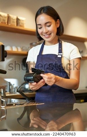 Beautiful smiling asian girl, barista insert client credit card in POS terminal, processing payment, taking orders in coffee shop.
