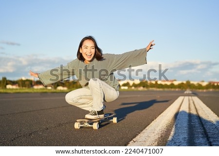 Freedom and happiness. Happy asian girl riding her longboard on an empty sunny road, laughing and smiling, skateboarding. Royalty-Free Stock Photo #2240471007