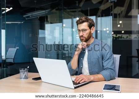 .Serious pensive businessman in shirt thinking about decision sitting at table in modern office, man with beard is using laptop at work. Royalty-Free Stock Photo #2240486645