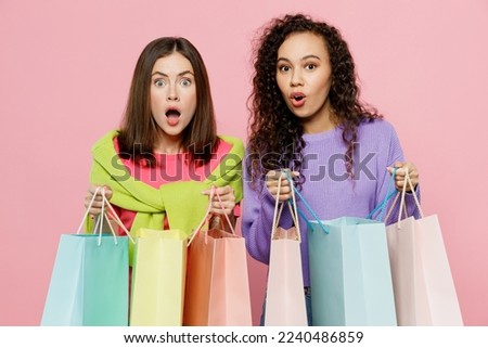 Young two friends exultant shocked women wears shirts hat hold in hand open paper package bags after shopping together isolated on plain light pink color background. Black Friday sale buy day concept