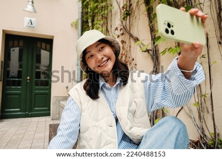 Smiling cute asian girl wearing panama taking selfie with mobile phone while sitting outdoors at city street