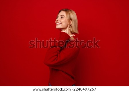 Smiling blonde woman in red sweater pointing finger at copy space isolated over red studio wall