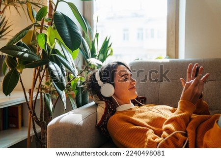 Young white woman in headphones using cellphone while lying on couch at home Royalty-Free Stock Photo #2240498081