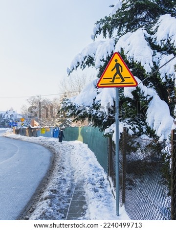 Pedestrian crossing road sign on a background of snow-covered trees, a footpath and a highway on a frosty winter day. Heavy snowfall, seasonal danger for pedestrians and drivers