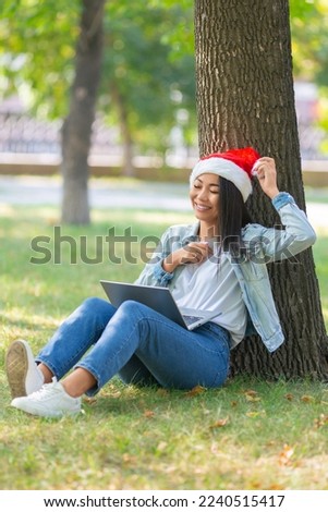 Vertical photo of a young woman with a laptop in the park. She sits on the grass in Santa Hat and communicates with relatives on the eve of Christmas. Online video call. Modern technology cincept.