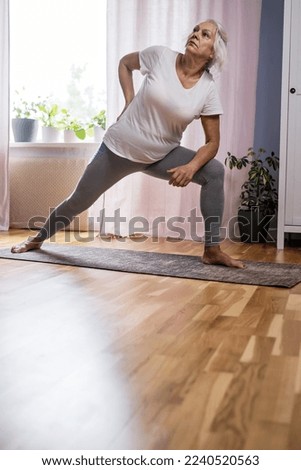 Domestic yoga practice. Positive mature lady standing in yoga pose, keeping balanced during covid lockdown at home. Happy senior lady in sportswear exercising 