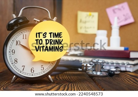 Medical concept. On the alarm clock a sticker with the inscription - It's time to drink vitamins. In the background, out of focus, there is a stethoscope, a nasal spray, a thermometer and a notepad.