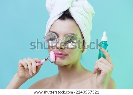  Horizontal photo, a woman with radiant skin on a blue background in a towel on her head and body and iridescent glasses and patches on her lips takes care of her face with serum