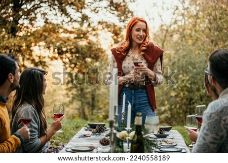 Attractive redhead woman standing by the table during a garden party and announcing and making a toast with a glass of wine with her friends. Royalty-Free Stock Photo #2240558189