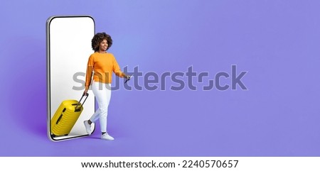 Cool pretty young black woman traveller walking from big cell phone with white empty screen, carrying yellow luggage, holding passport with tickets, mockup, purple studio background, copy space