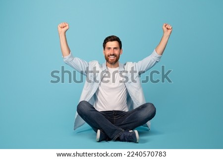 Smiling excited young european male raises his hands up and makes sign of victory and success, sits on floor in lotus position, isolated on blue background, studio. Emotions, reactions to great news