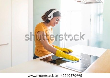 Happy Middle Eastern Female Listening Music In Headphones While Cleaning Kitchen, Cheerful Young Arab Woman Polishing Induction Cooktop Surface With Rag And Enjoying Favorite Songs, Copy Space Royalty-Free Stock Photo #2240570995