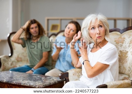 Dissatisfied mother-in-law siting on a chair against of a confused husband and wife on the sofa in the living room at home Royalty-Free Stock Photo #2240577223