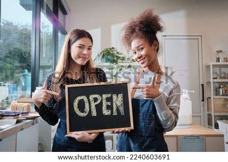 Two young female shopkeepers show open sign board with cheerful smiles in refill store shop, happy work with organic products, zero waste groceries, eco-friendly merchandise, and sustainable business.