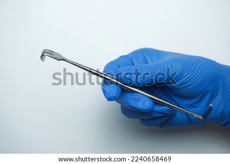 Doctor wearing blue gloves holding sharp Senn-Miller Retractor is a multi-purpose, dual-ended traction device that can be used to open superficial wounds in common surgical procedures