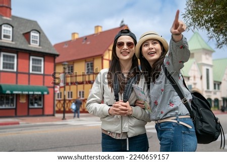 travel lifestyle and people on vacation in usa. two smiling asian girl backpackers pointing at famous attraction in space near colorful Danish houses at background in solvang