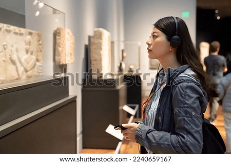 side view of asian Korean college girl taking audio tour in the history museum located in los angeles usa. she is looking at an ancient carving displayed in glass case Royalty-Free Stock Photo #2240659167