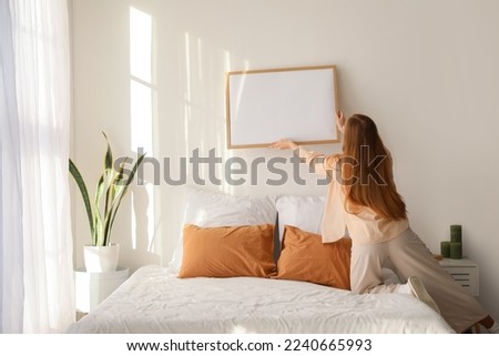 Young woman hanging blank frame on light wall in bedroom, back view Royalty-Free Stock Photo #2240665993