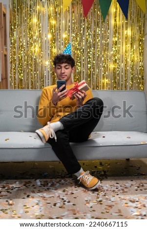 Happy pretty arabic guy in festive hat typing text on smartphone, chatting online, reading greeting messages, celebrating birthday party at home, making video call to friend, family. Copy space