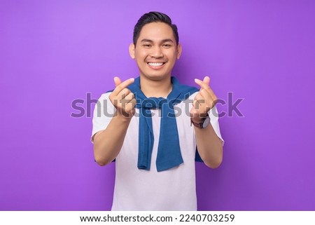 Smiling young handsome and stylish Asian man wearing white t-shirt casual clothes showing Korean heart love sign isolated on purple background. People lifestyle concept Royalty-Free Stock Photo #2240703259