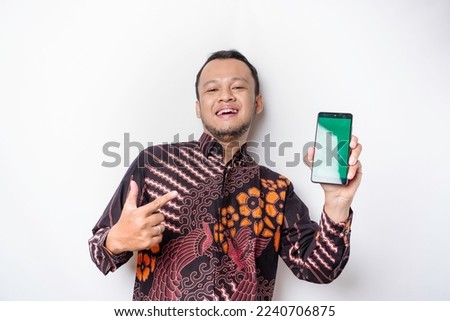 A portrait of a smiling Asian man wearing a batik shirt and showing green screen on her phone, isolated by white background