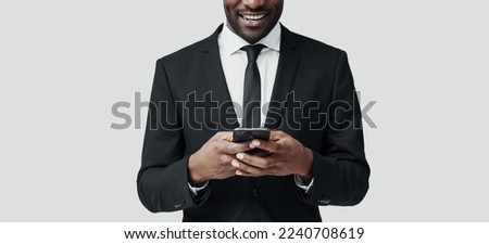Modern young African man in formalwear using smart phone and smiling while standing against grey background