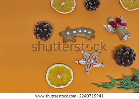 Christmas gingerbread in the shape of a star, straw ornaments in the shape of an angel and a star, pine cones, orange slices and a green twig on an orange background