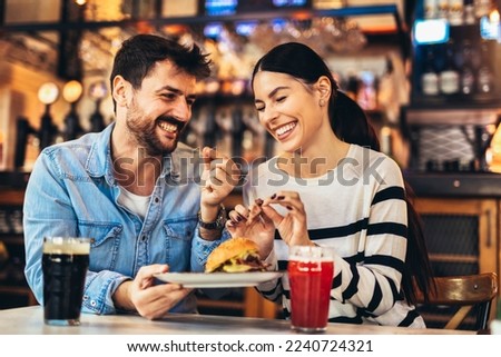 Young couple in love having fun spending leisure time together at restaurant, eating burgers and drinking beer Royalty-Free Stock Photo #2240724321