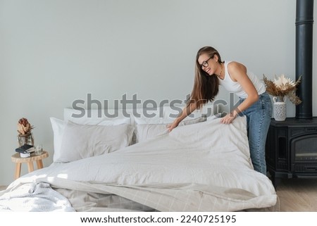 Laughing caucasian beautiful young woman makes bed wears jeans and white t-shirt and glasses. Pretty hispanic girl makes housekeeping. Mockup, people at home. Cheerful student makes order home.