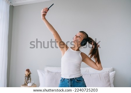 Fit healthy Caucasian young woman in white t-shirt and blue jeans uses headphones makes selfie at new home. Pretty hispanic girl makes video cal from hotel room enjoys vacations. Youth and travel.