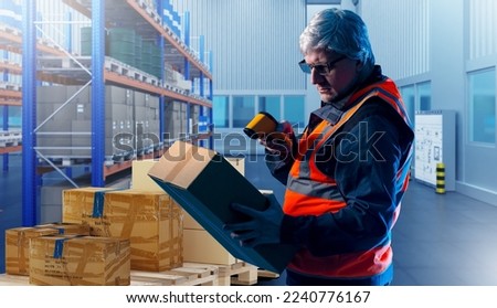 Warehouse worker. Man storekeeper with device for revision. Gray-haired man works in warehouse. Inspector checks box using data collection terminal. Storekeeper expressed vest in in warehouse hangar