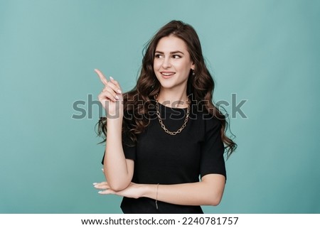 Adorable Italian brunette girl  smiles, points by index fingers aside at empty space on turquoise studio background. Attractive hispanic young woman recommends something. Mockup, sale, discount.