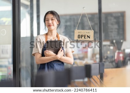 Female Asian coffee shop small business owner wearing apron standing in open sign front of counter performing stock check. afro hair employee Barista entrepreneur. 