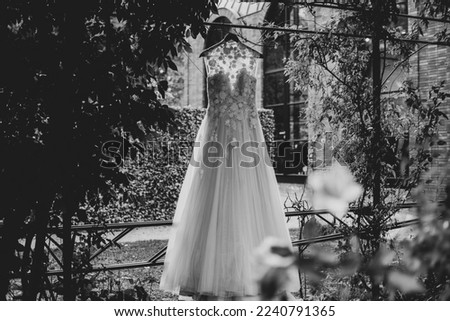 Beautiful long classic wedding dress in black and white on a hanger in the open air between the green climbing plants at wedding venue. 