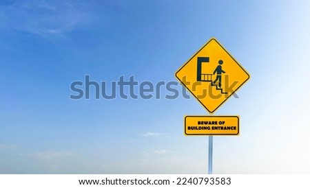 Yellow sign with graphic of person stepping down stairs and the text beware of building entrance on blue sky background