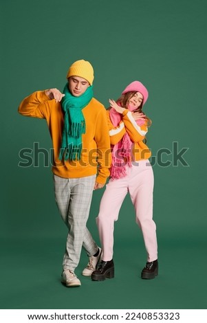 Portrait of young man and woman in bright knitted hat and scarf posing, dancing isolated over green background. Concept of emotions, winter holidays, fashion, lifestyle, celebration, relationship