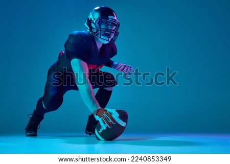 Start. Sportive, strong man, american football player in sports team uniform and protective helmet isolated over blue background in neon light. Power, energy, achievements, skills and ad