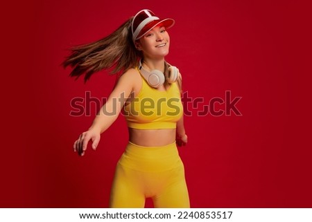 Portrait of young woman in yellow sportswear posing in plastic cap, training isolated over red background. Concept of youth culture, emotions, facial expression, sport, lifestyle, health