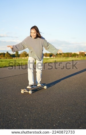 Cute asian girl riding skateboard, skating on road and smiling. Skater on cruiser longboard enjoying outdoors on sunny day.