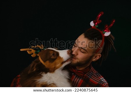 Concept of pet celebrating Merry Christmas with male owner. Young Caucasian man with beard and dreadlocks hugs Australian Shepherd and kisses smiling. Studio shot. Dog and man with deer horns.
