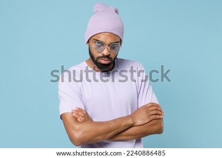 Young sad tired serious shrewd unshaven man of African American ethnicity he wear violet t-shirt hat hold hands crossed folded isolated on plain pastel light blue cyan color background studio portrait